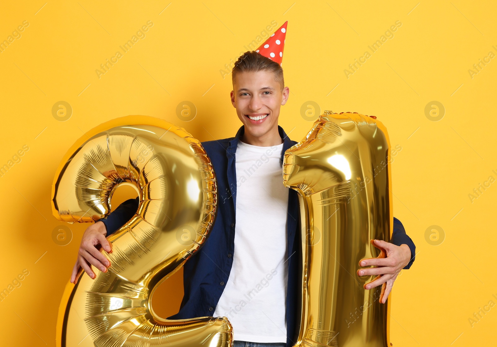 Photo of Coming of age party - 21st birthday. Happy young man with number shaped balloons and hat on yellow background