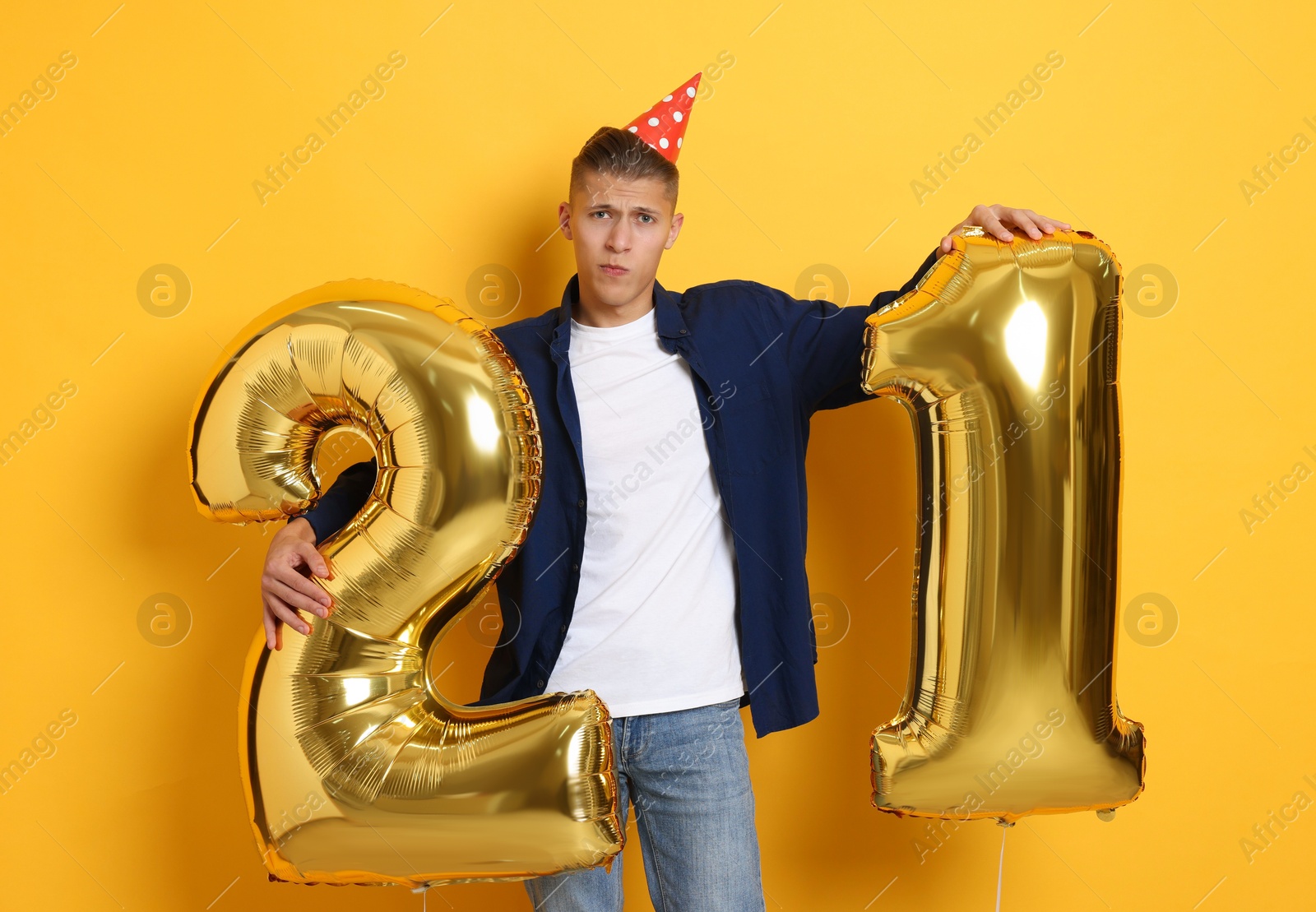 Photo of Coming of age party - 21st birthday. Upset young man with number shaped balloons and hat on yellow background