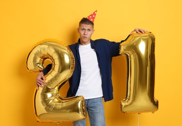 Photo of Coming of age party - 21st birthday. Upset young man with number shaped balloons and hat on yellow background