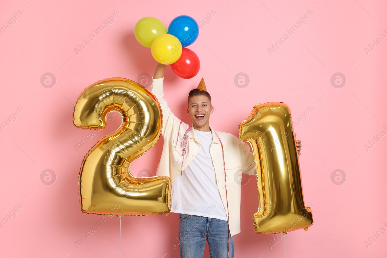 Photo of Coming of age party - 21st birthday. Happy young man with number shaped balloons and hat on pink background