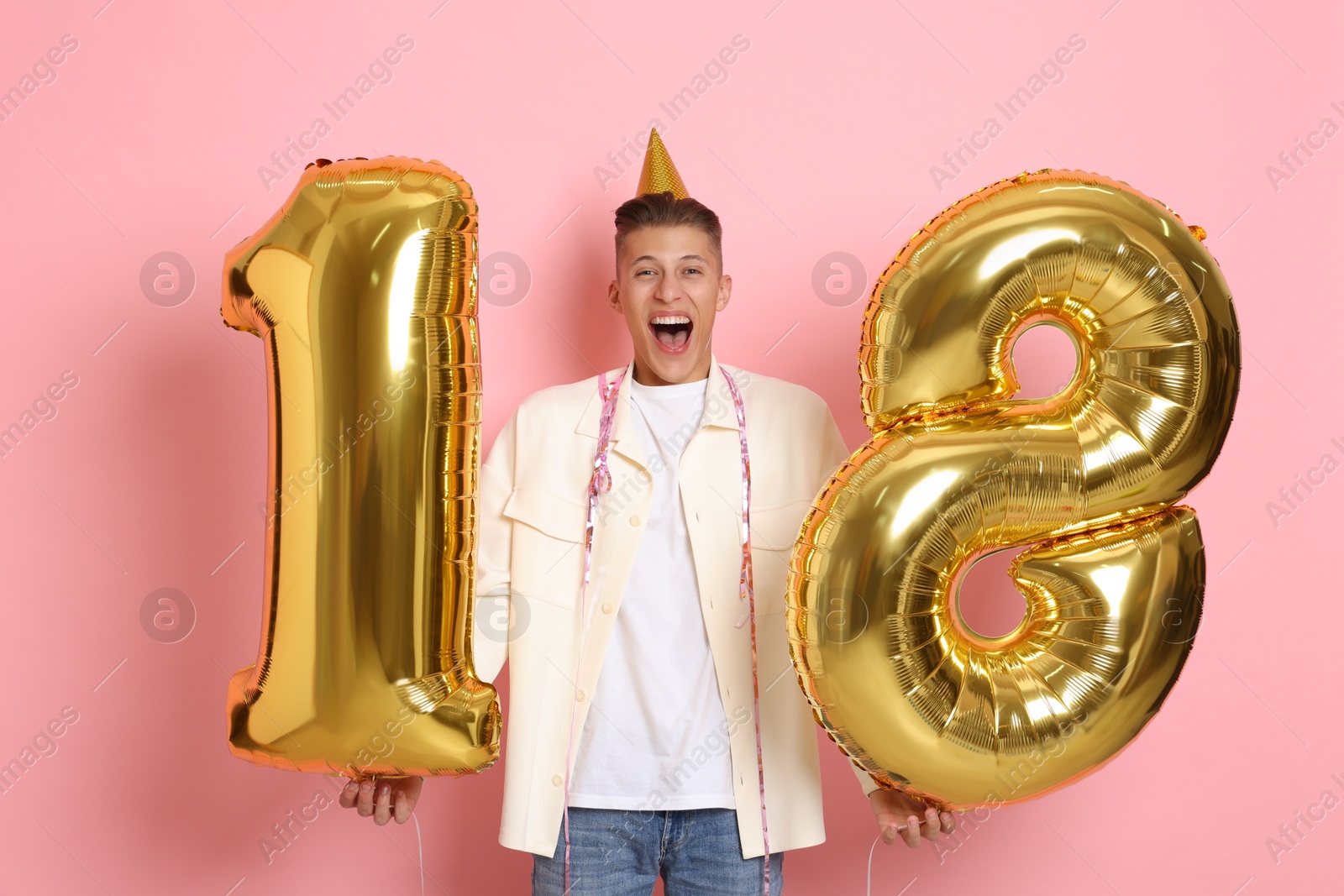 Photo of Coming of age party - 18th birthday. Emotional young man with number shaped balloons and hat on pink background