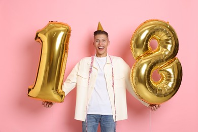 Photo of Coming of age party - 18th birthday. Happy young man with number shaped balloons and hat on pink background