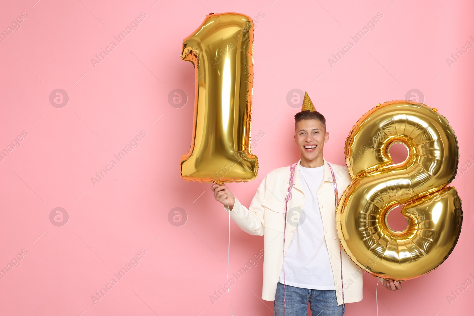 Photo of Coming of age party - 18th birthday. Happy young man with number shaped balloons and hat on pink background, space for text