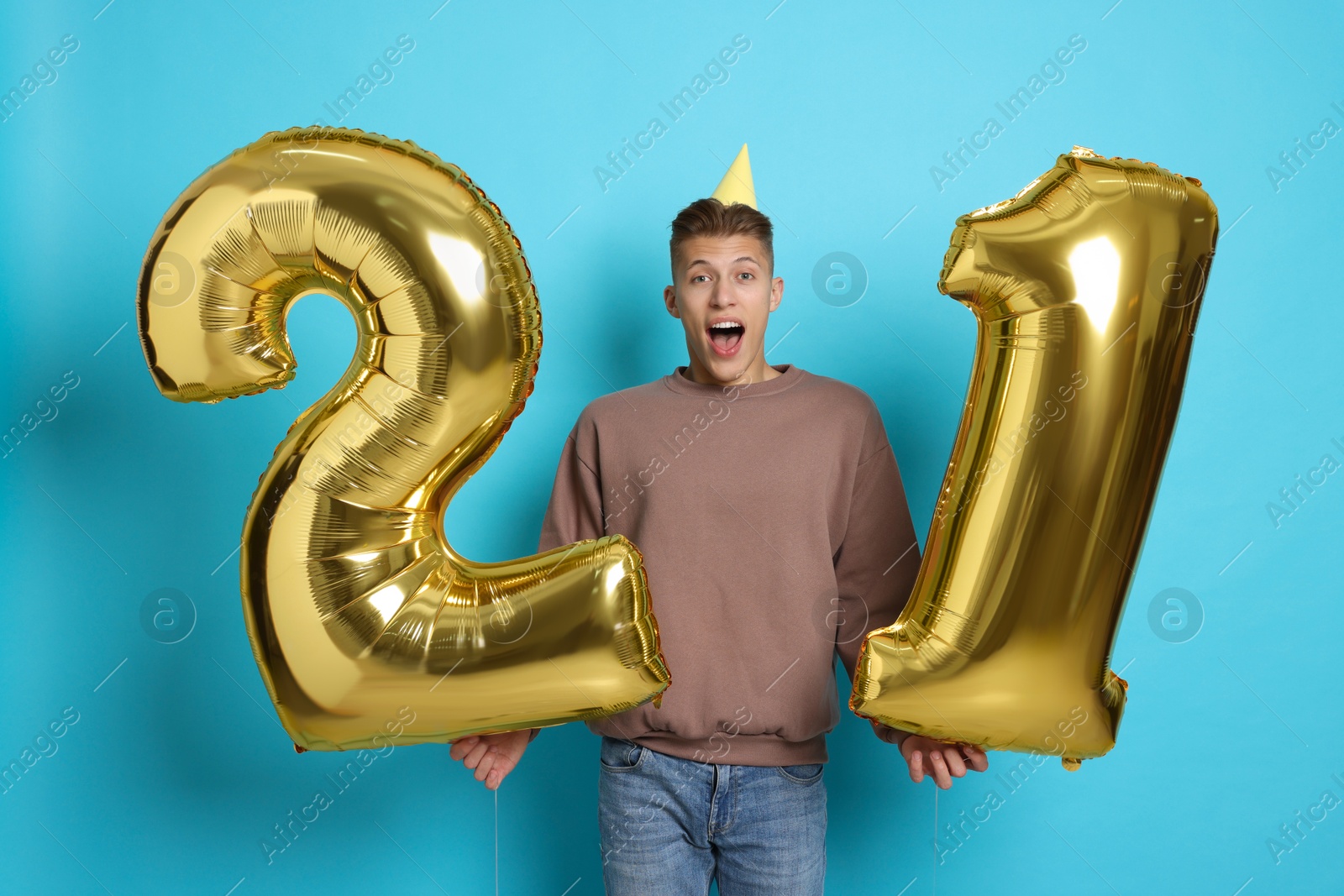 Photo of Coming of age party - 21st birthday. Emotional young man with number shaped balloons and hat on light blue background