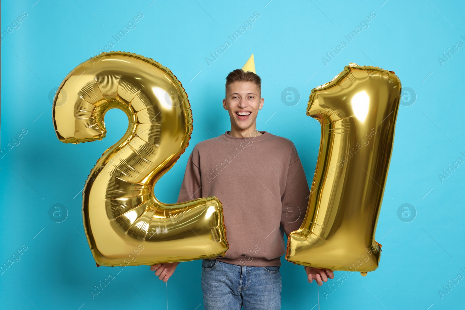 Photo of Coming of age party - 21st birthday. Happy young man with number shaped balloons and hat on light blue background