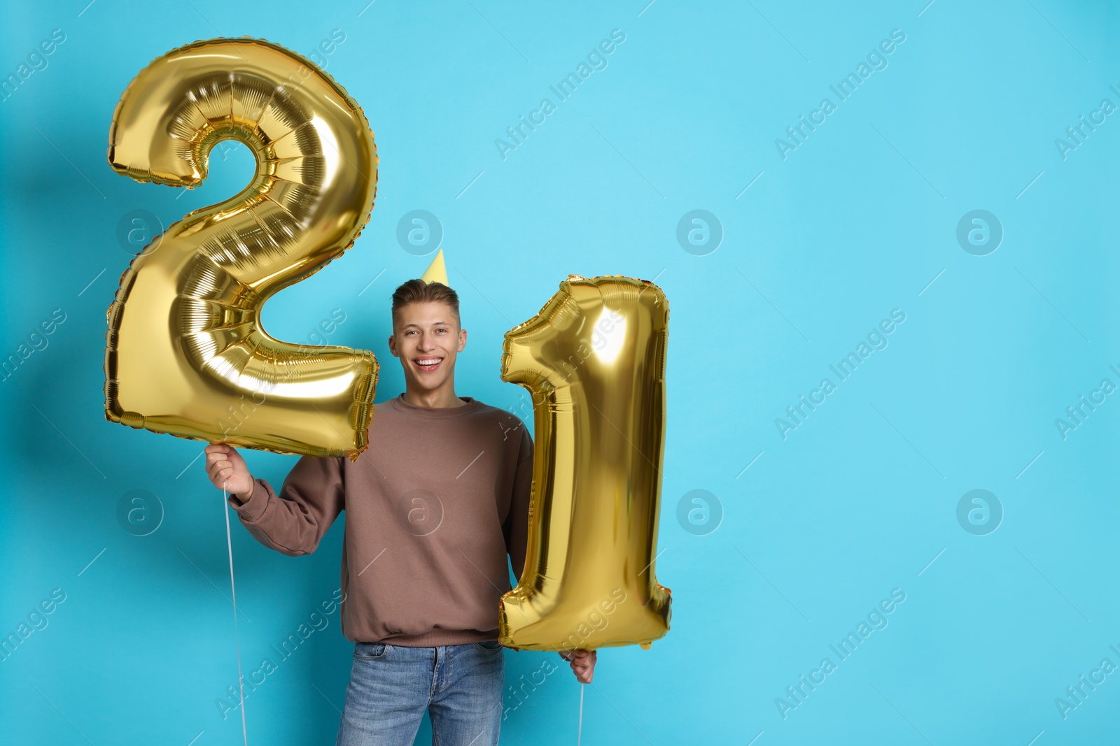 Photo of Coming of age party - 21st birthday. Happy young man with number shaped balloons and hat on light blue background, space for text