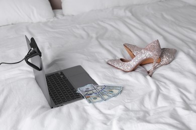 Photo of Prostitution and virtual sex. Laptop with web camera, dollar banknotes and high heels on bed