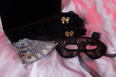 Photo of Prostitution and virtual sex. Laptop, dollar banknotes and mask on linens