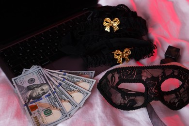 Photo of Prostitution and virtual sex. Laptop, dollar banknotes and mask on linens