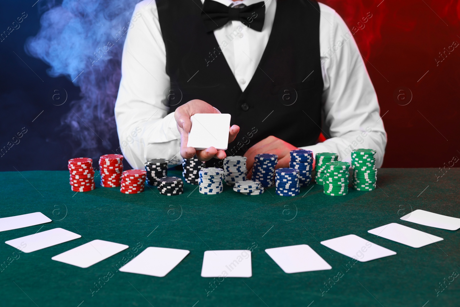 Photo of Professional croupier holding playing cards and casino chips at gambling table against color background with smoke, closeup