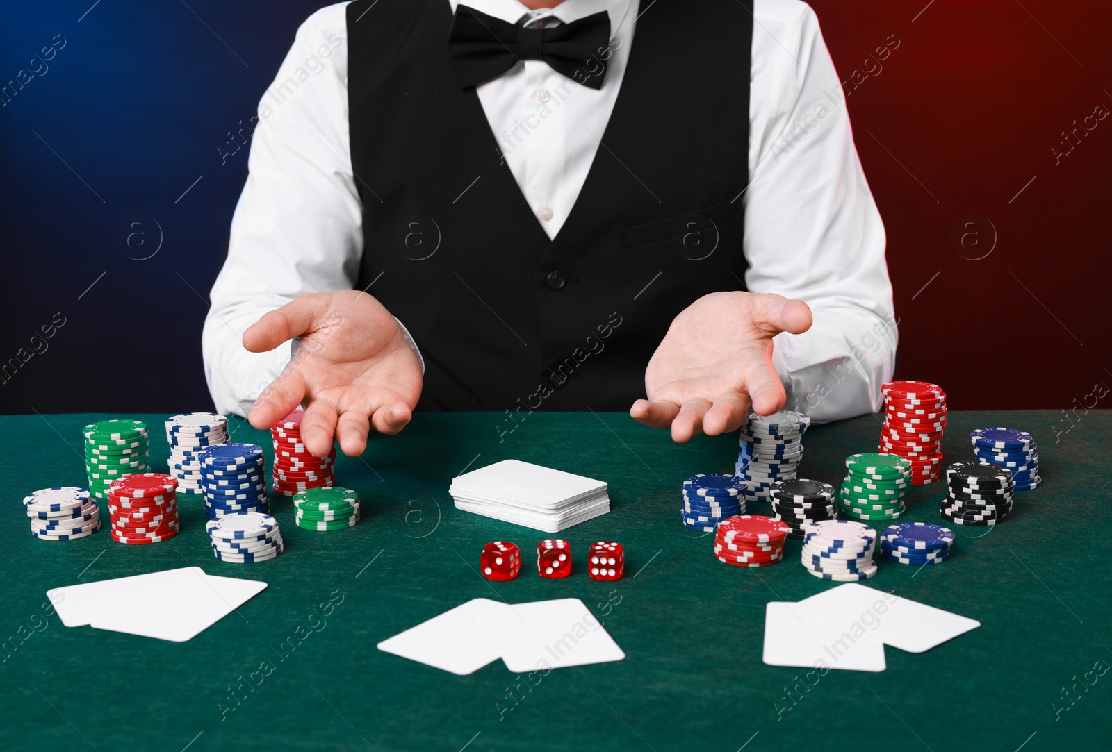 Photo of Professional croupier at gambling table with playing cards, casino chips and dice against color background, closeup