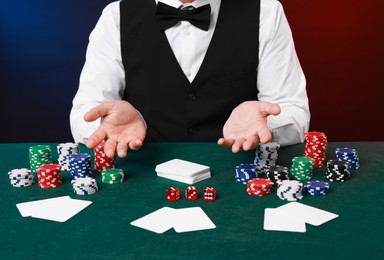 Professional croupier at gambling table with playing cards, casino chips and dice against color background, closeup