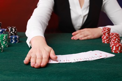 Photo of Professional croupier with casino chips and playing cards at gambling table on color background, closeup