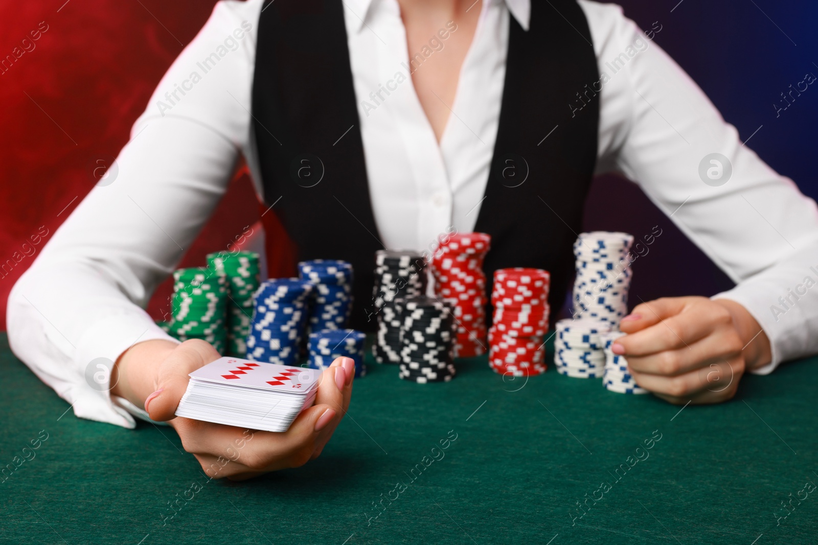 Photo of Professional croupier with casino chips and playing cards at gambling table on color background with smoke, closeup