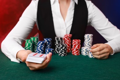 Photo of Professional croupier with casino chips and playing cards at gambling table on color background with smoke, closeup