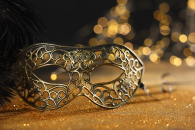 Photo of Beautiful carnival mask and black feathers on golden background against blurred lights, closeup. Bokeh effect