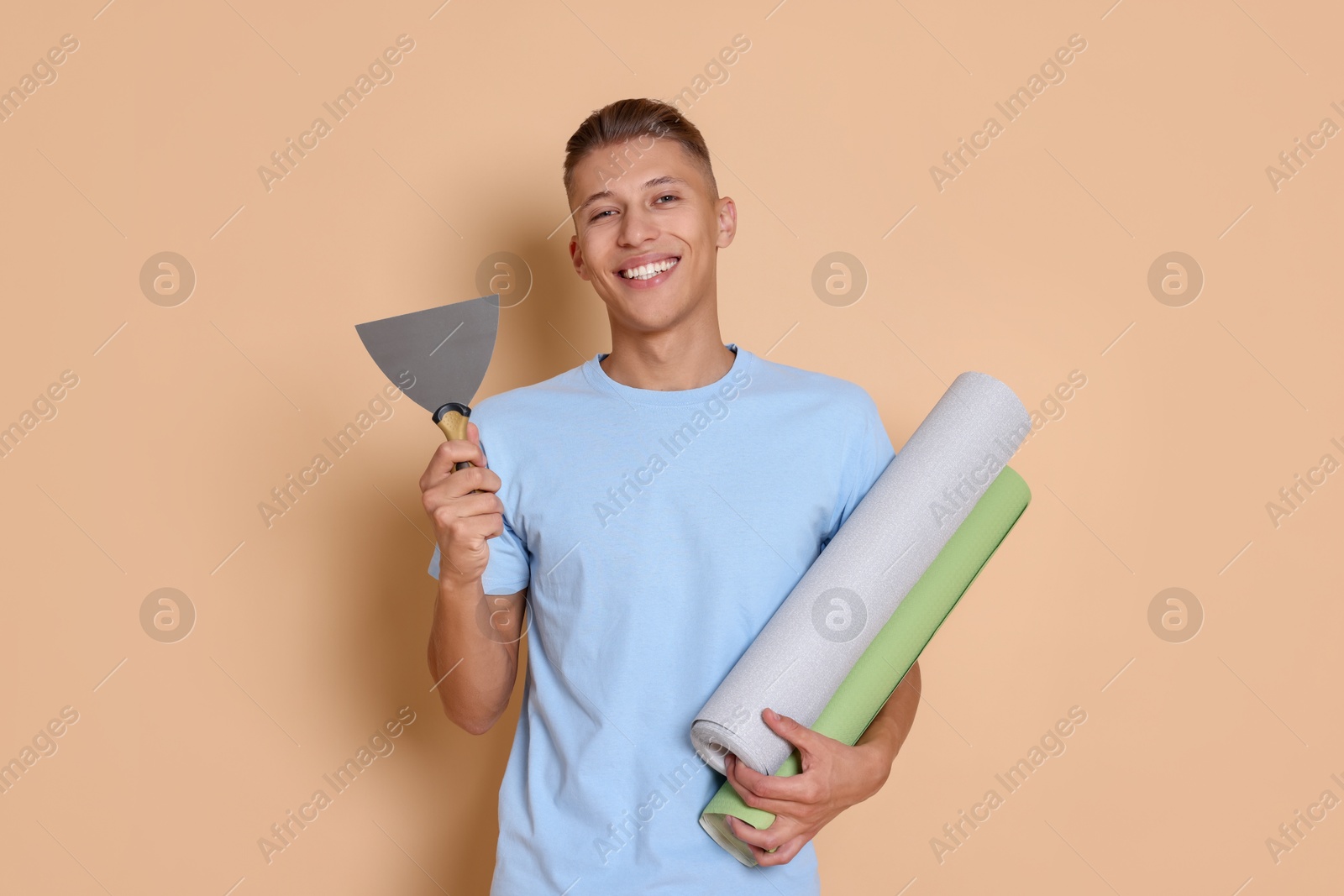 Photo of Young decorator with putty knife and rolls of wallpaper on beige background