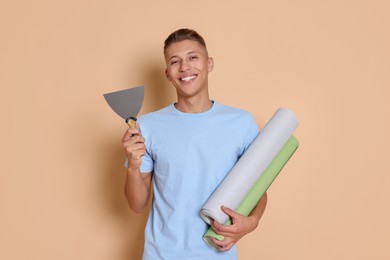 Photo of Young decorator with putty knife and rolls of wallpaper on beige background