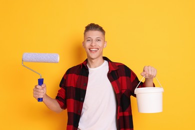 Photo of Portrait of young decorator with paint roller and bucket on orange background