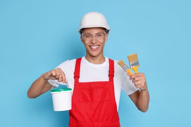 Photo of Portrait of young decorator with bucket and brushes on light blue background