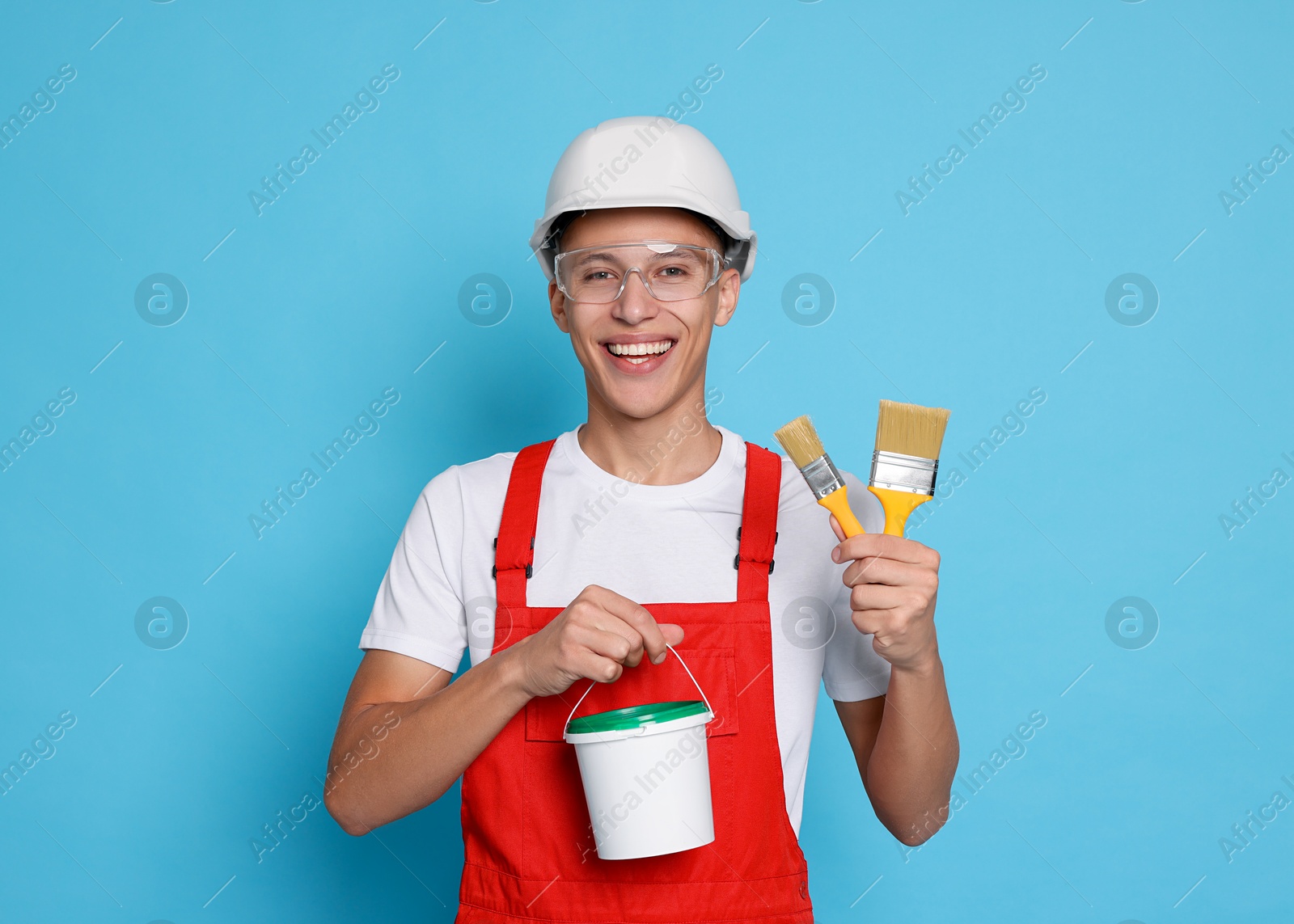 Photo of Portrait of young decorator with bucket and brushes on light blue background