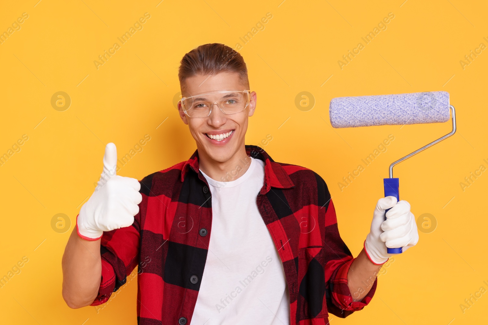 Photo of Portrait of young decorator with paint roller showing thumbs up on orange background