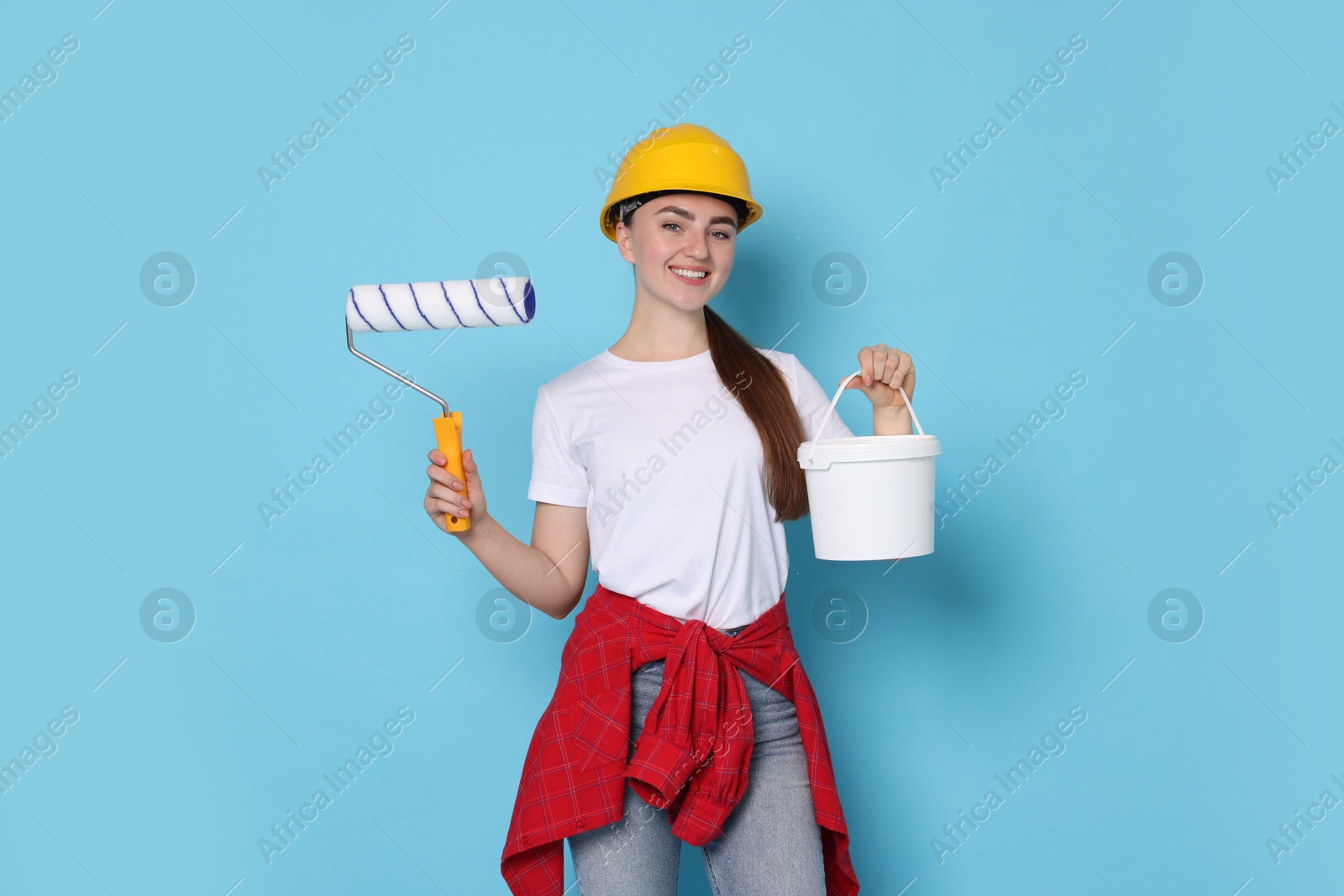 Photo of Portrait of young decorator with paint roller and bucket on light blue background