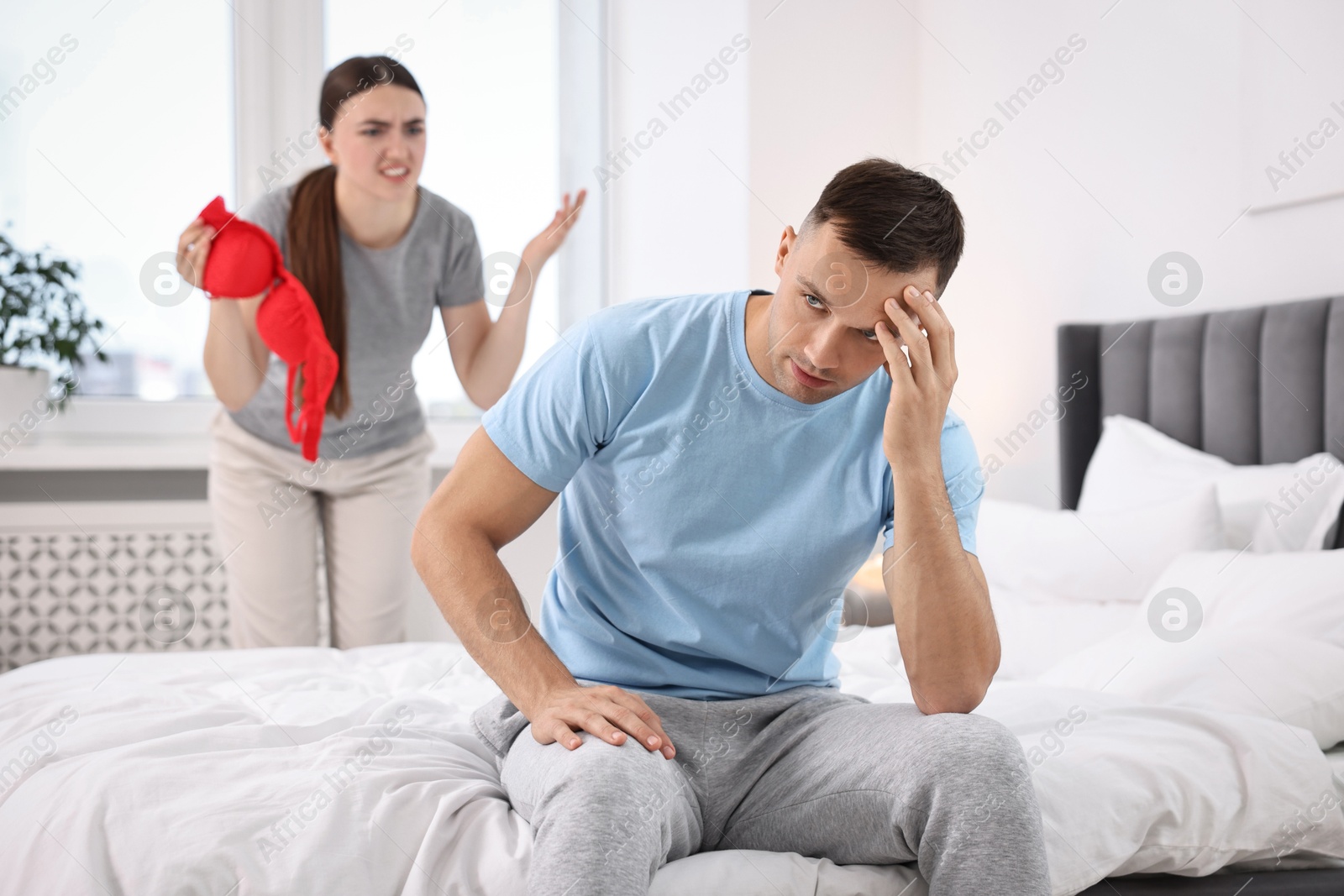 Photo of Angry wife showing mistress bra as proof to her unfaithful husband while he ignoring her in bedroom, selective focus