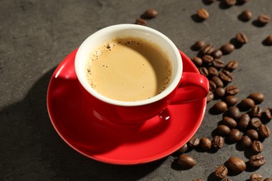 Photo of Red cup of aromatic coffee, saucer and beans on dark textured table, closeup