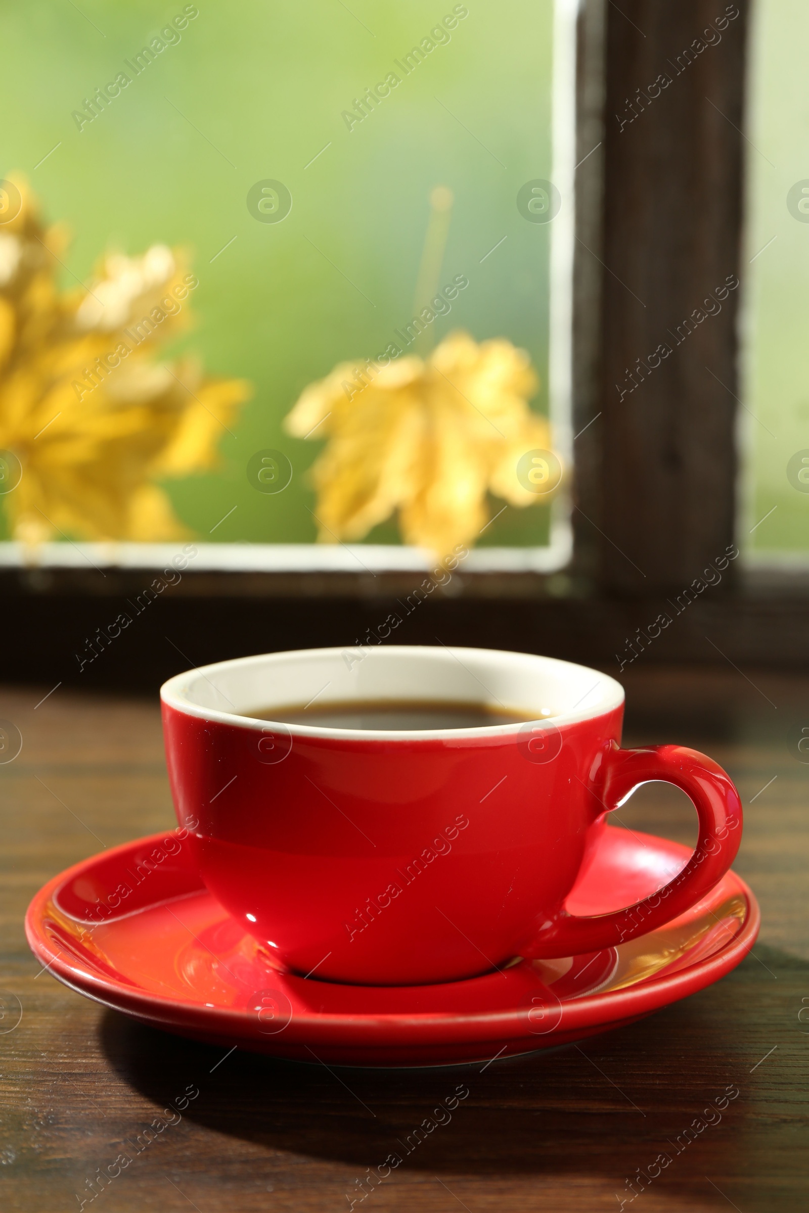 Photo of Red cup of aromatic coffee and saucer on wooden sill near window