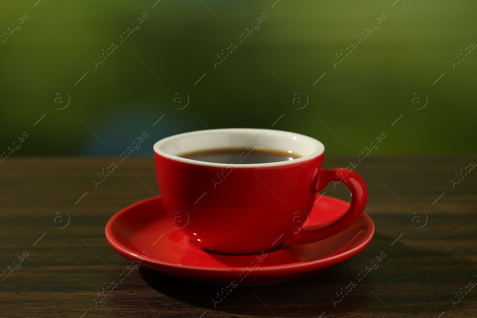 Photo of Red cup of aromatic coffee and saucer on wooden table against blurred green background, closeup