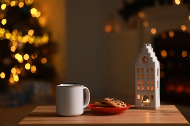 Photo of Mug of drink, cookies and decorative candle holder on wooden table in room with Christmas lights