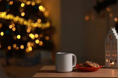 Photo of Mug of drink, cookies and decorative candle holder on wooden table in room with Christmas lights