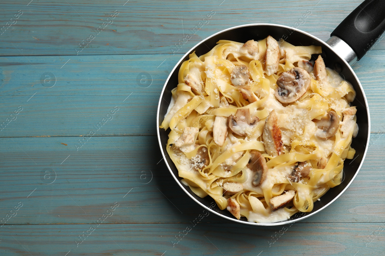 Photo of Tasty Alfredo pasta with chicken and mushrooms in frying pan on light blue wooden table, top view. Space for text