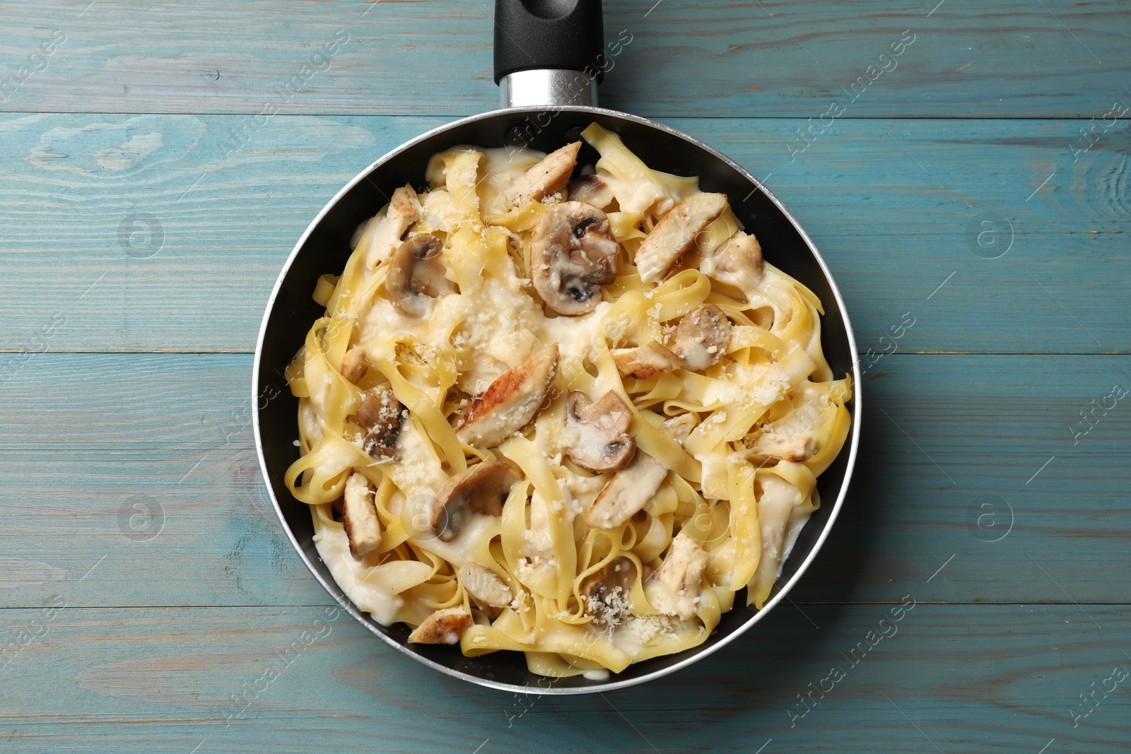 Photo of Tasty Alfredo pasta with chicken and mushrooms in frying pan on light blue wooden table, top view