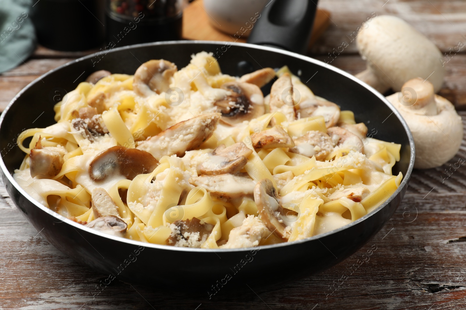 Photo of Tasty Alfredo pasta with chicken and mushrooms in frying pan on wooden table, closeup