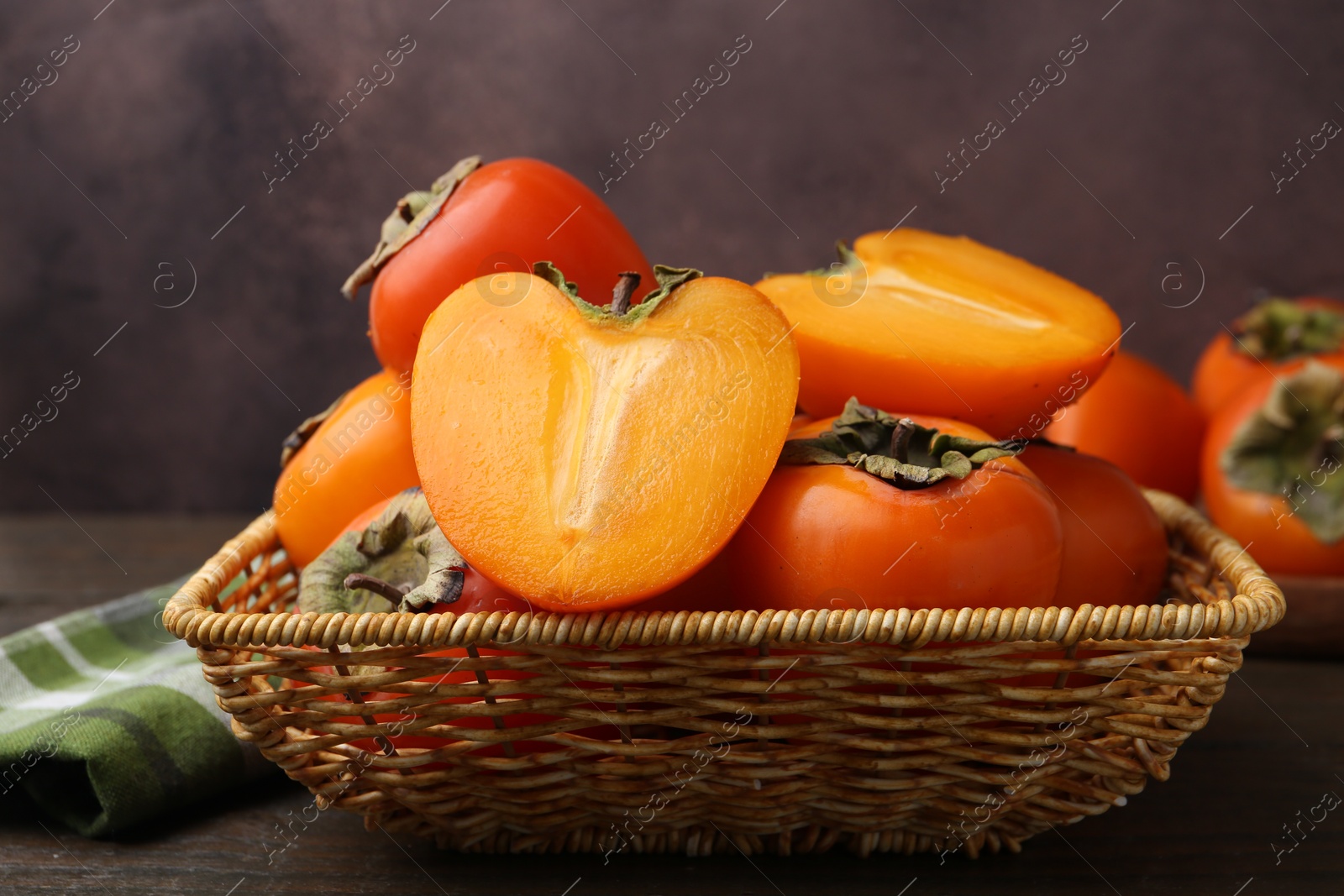 Photo of Whole and cut juicy persimmons in wicker basket on wooden table, closeup