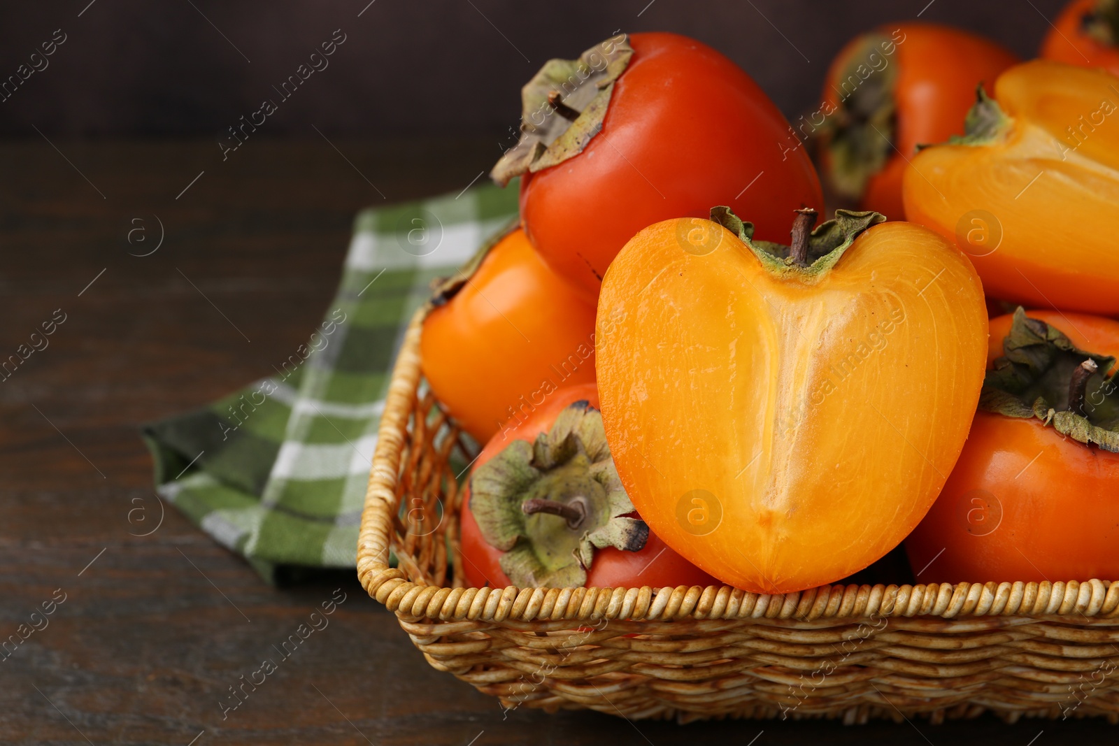 Photo of Whole and cut juicy persimmons in wicker basket on wooden table, closeup. Space for text