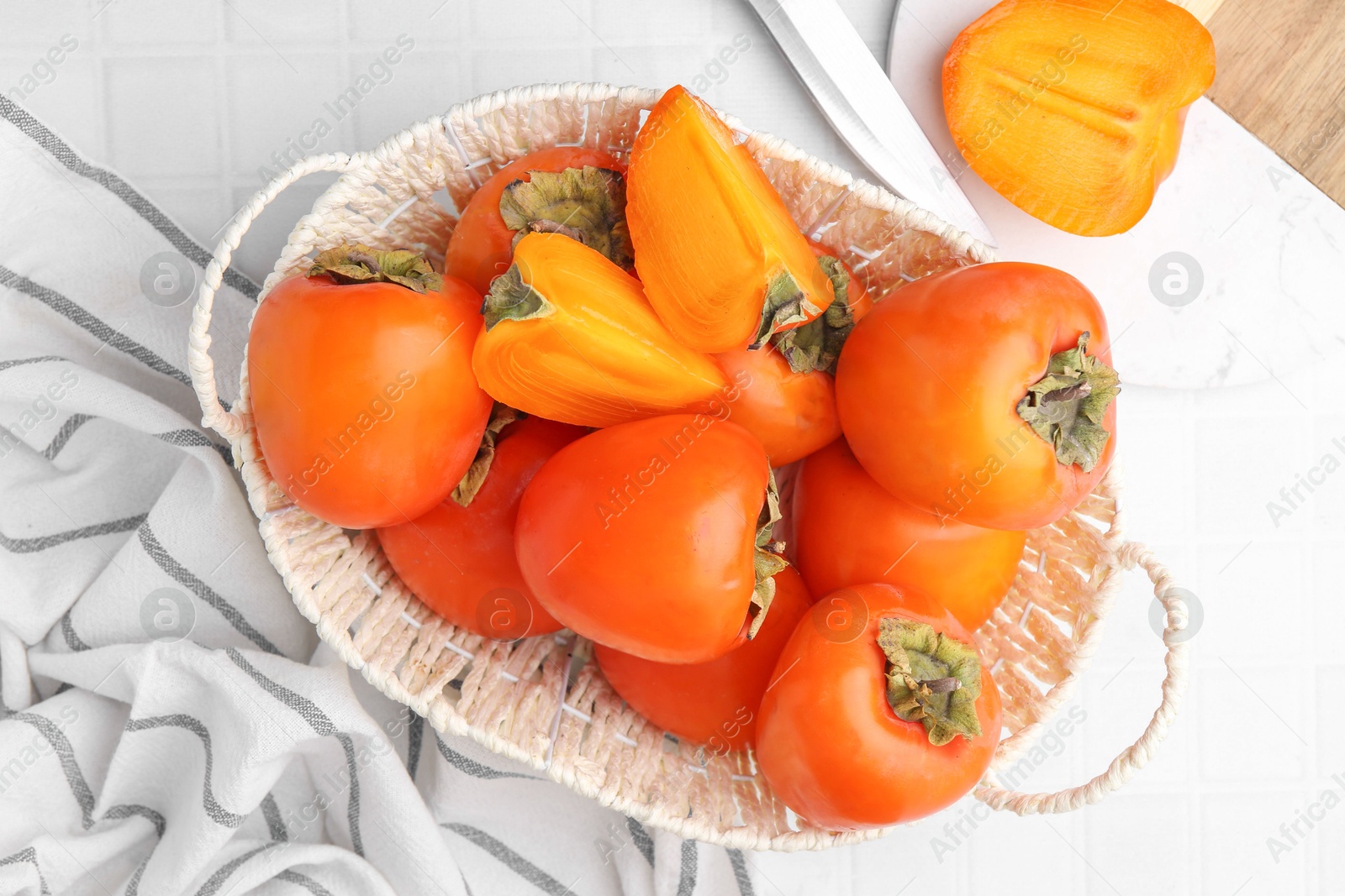 Photo of Ripe persimmons in wicker basket on white tiled table, top view