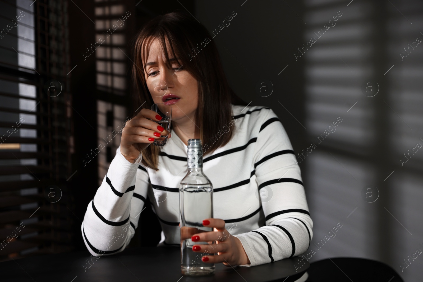 Photo of Alcohol addiction. Woman with shot glass of vodka and bottle at table indoors