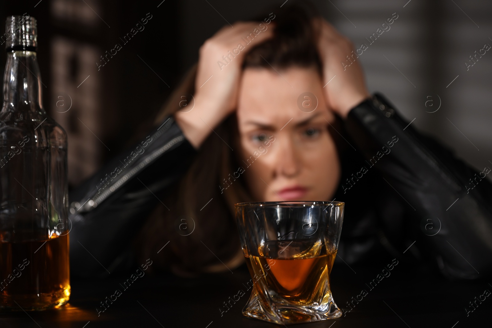 Photo of Alcohol addiction. Woman at table indoors, focus on glass of whiskey and bottle