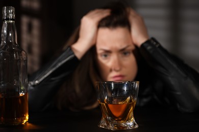 Alcohol addiction. Woman at table indoors, focus on glass of whiskey and bottle