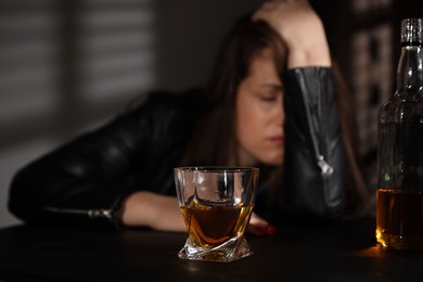 Photo of Alcohol addiction. Woman at table indoors, focus on glass of whiskey and bottle