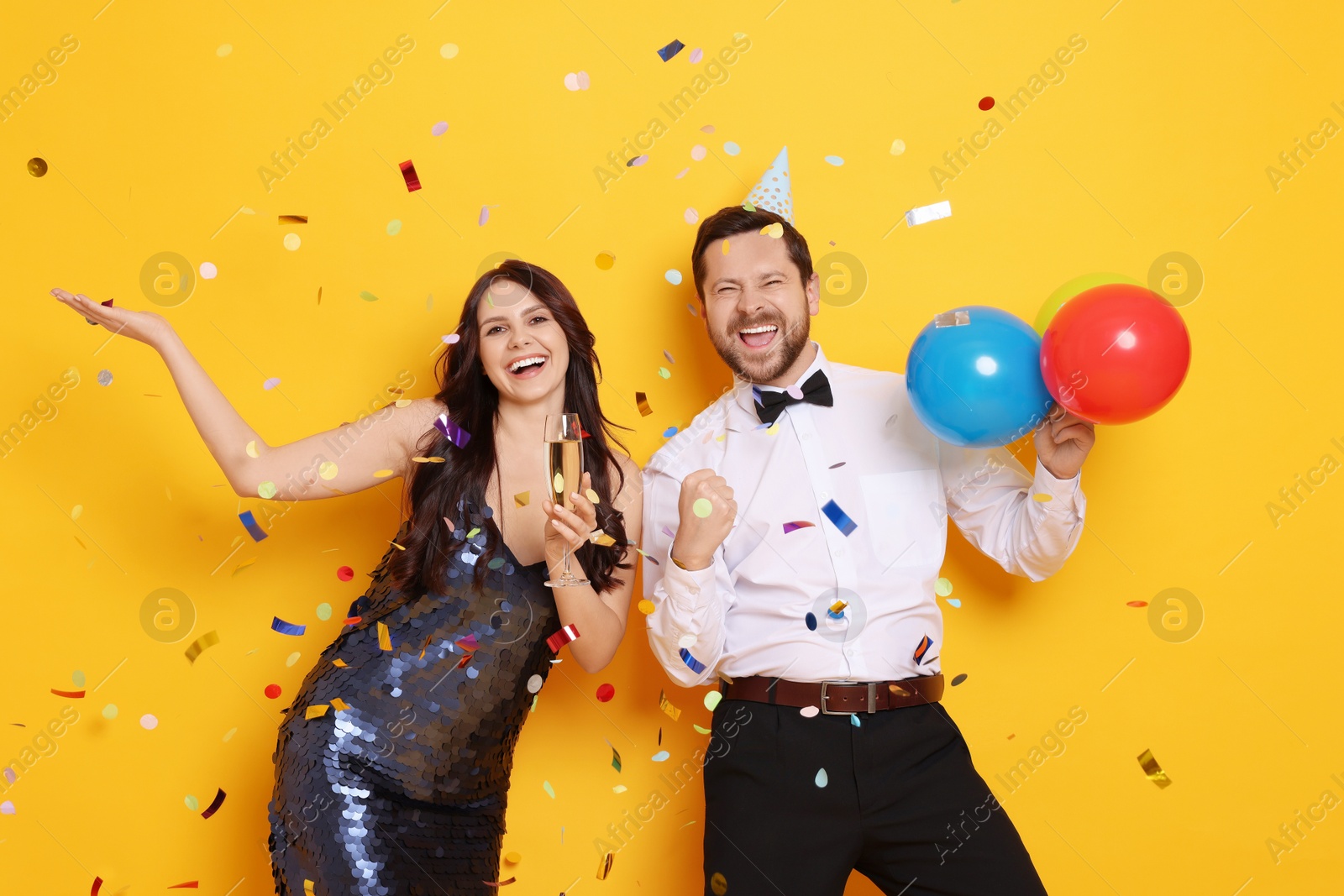 Photo of Happy friends with sparkling wine and balloons under falling confetti on orange background. Surprise party