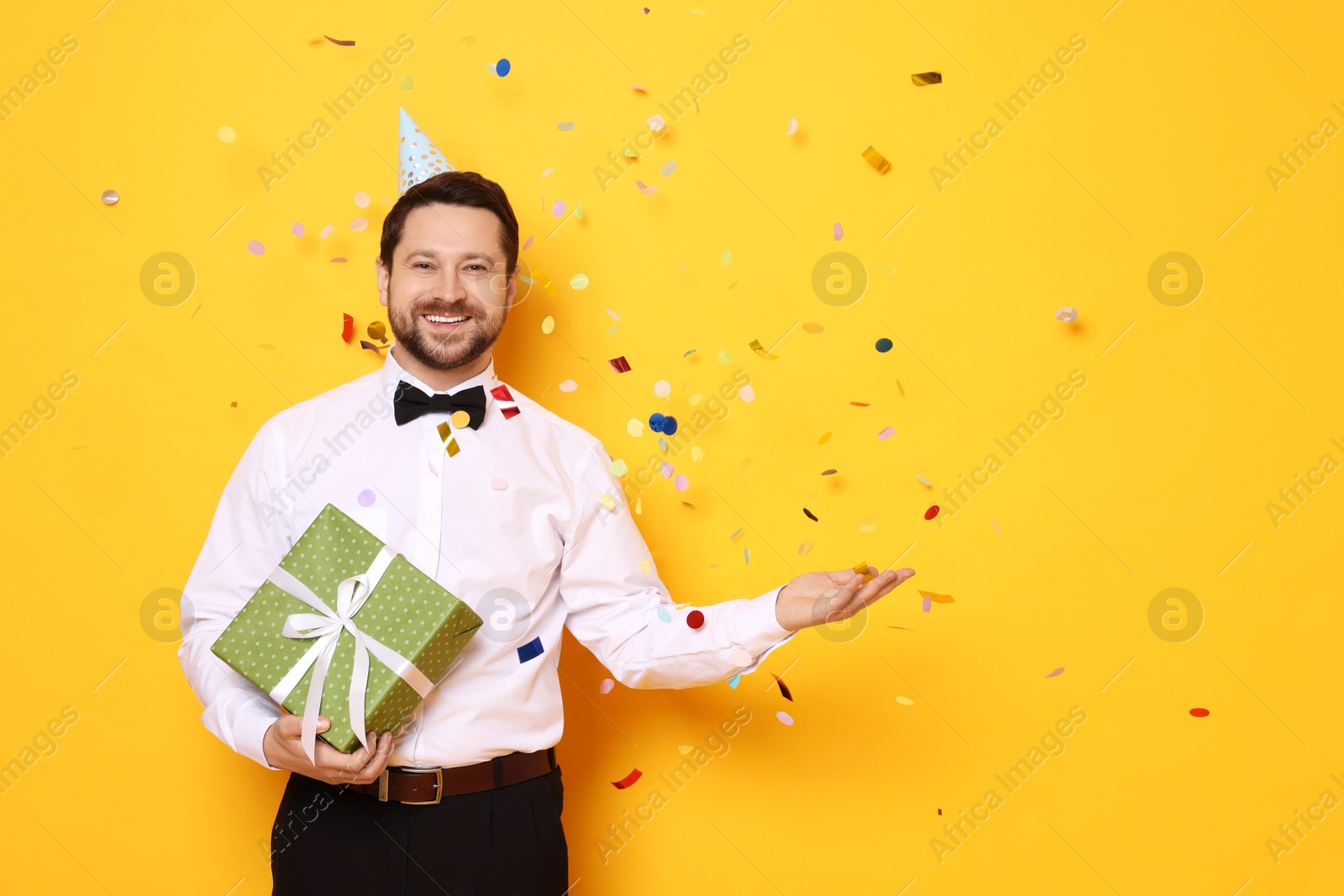 Photo of Happy man with gift box under falling confetti on orange background, space for text. Surprise party