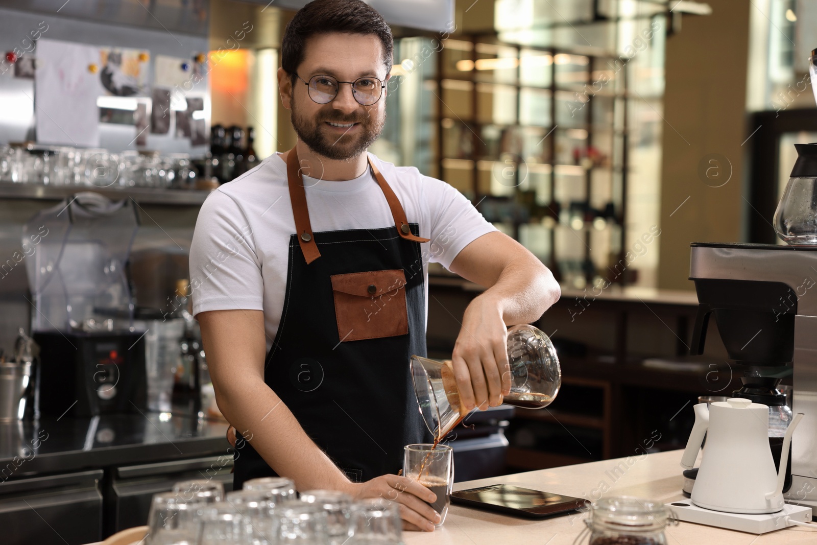 Photo of Barista pouring coffee from glass coffeemaker into cup at table in cafe