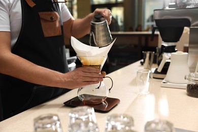 Photo of Barista brewing coffee in glass coffeemaker with paper filter at table in cafe, closeup