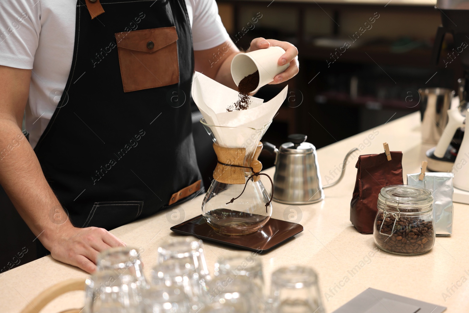 Photo of Barista pouring ground coffee into glass coffeemaker with paper filter at table in cafe, closeup