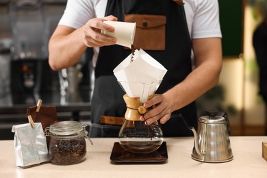 Photo of Barista pouring ground coffee into glass coffeemaker with paper filter at table in cafe, closeup