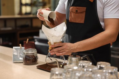 Barista pouring ground coffee into glass coffeemaker with paper filter at table in cafe, closeup